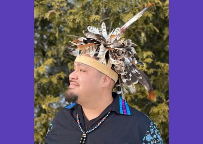 A person wearing a feathered headdress, black shirt with blue and purple patterned sleeves and a beaded necklace stands in front of green foliage. The photo appears over a plain purple background.