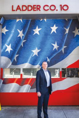 A man in a navy pinstripe suit stands in front of a fire station with "LADDER CO. 10" displayed above and a large American flag mural painted on the building's exterior wall.