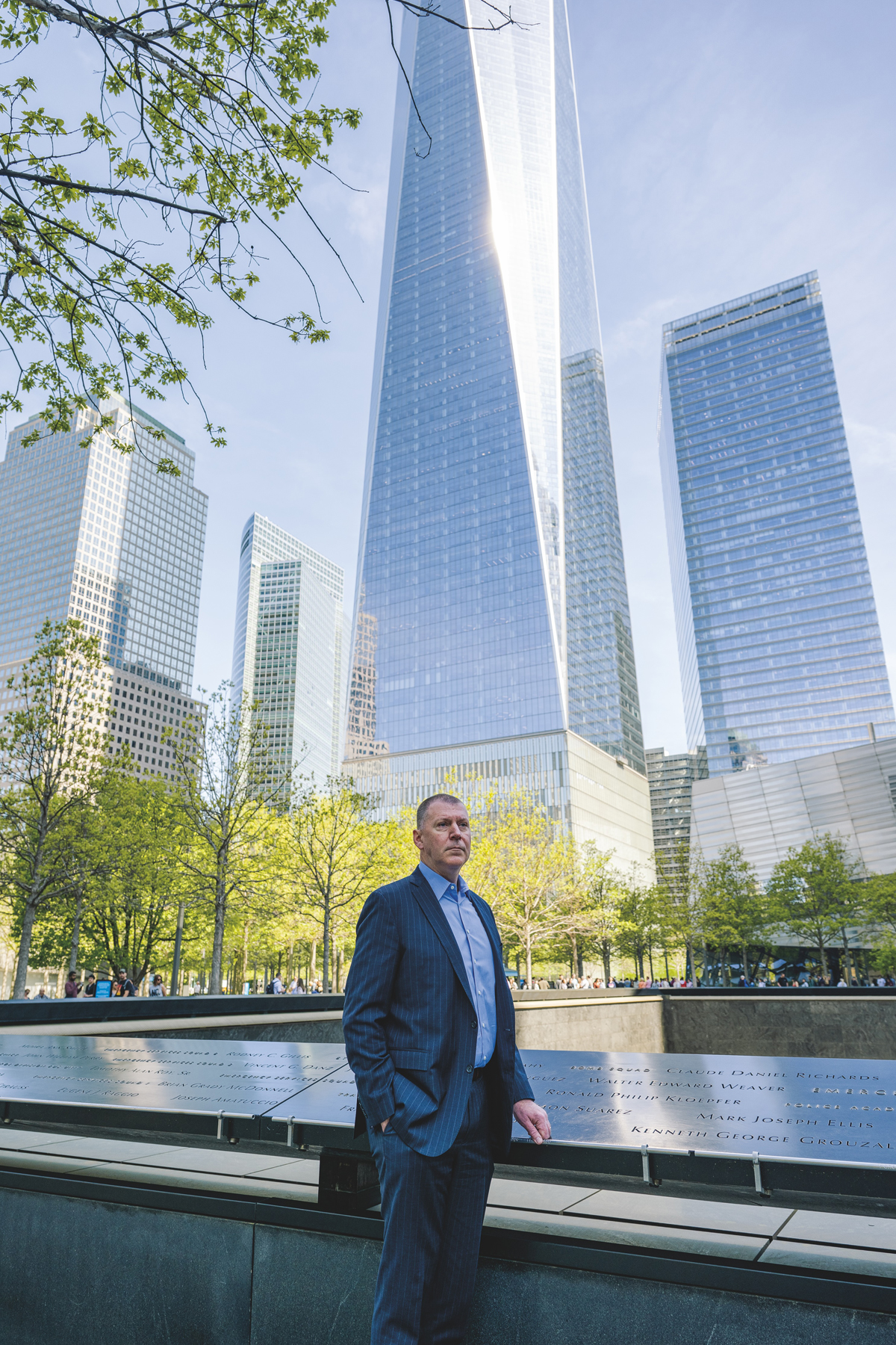 A man in a navy pinstripe suit and blue shirt stands at the 9/11 Memorial in New York City, with One World Trade Center and other skyscrapers towering in the background. The memorial's bronze panels with engraved names are visible, surrounded by spring trees in the memorial plaza.