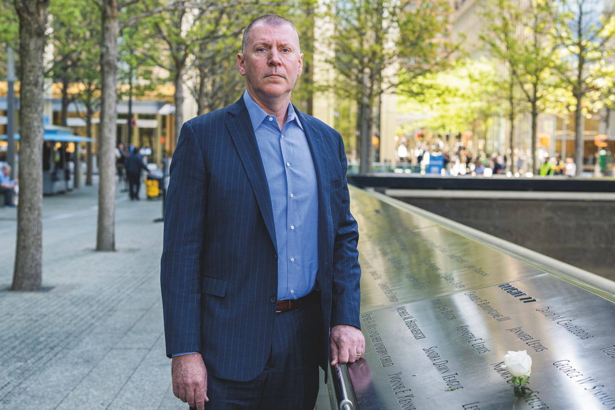 A man in a navy pinstripe suit and blue dress shirt stands solemnly beside the memorial panels at the 9/11 Memorial in New York City, with names of victims engraved in the reflective surface and a white flower placed among them. Trees and urban buildings are visible in the background.
