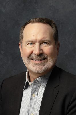 man with short hair and beard wearing a dark suit jacket over a light checkered shirt, posed against a textured dark gray background