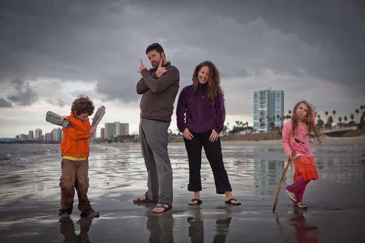 Four people posing playfully on a wet beach under a cloudy sky, with city buildings and palm trees in the background.