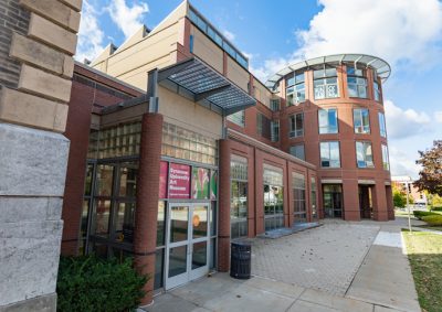 Modern brick and glass university museum building with curved cylindrical tower, glass entrance doors, and contemporary architectural design featuring red brick facades, large windows, and geometric elements under a partly cloudy blue sky.