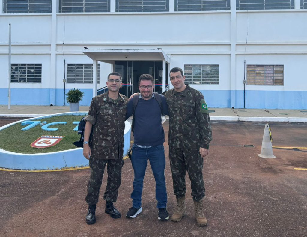 Three people standing in front of a blue and white building labeled '7C', including two in military uniforms and one in casual clothing