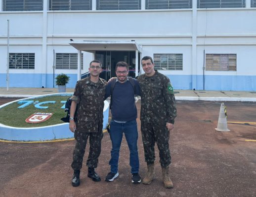 Three people standing in front of a blue and white building labeled '7C', including two in military uniforms and one in casual clothing.