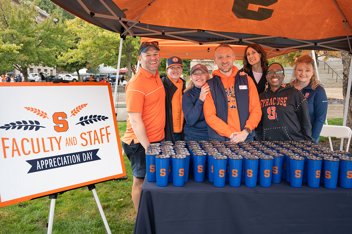 Group of adults in blue and orange gear pose behind a table of blue Syracuse branded tumblers at an outdoor Faculty and Staff Appreciation Day event under an orange tent