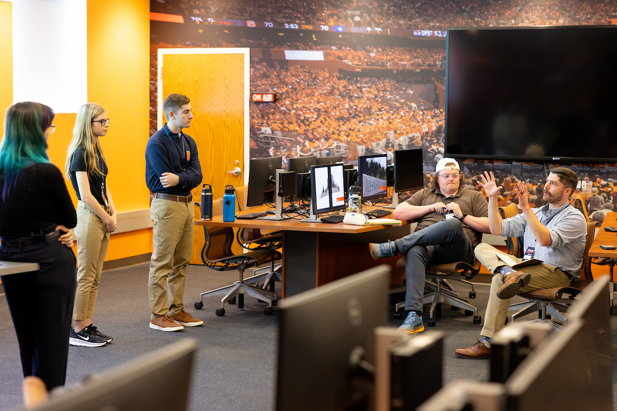 People in a broadcast studio with orange decor. Three stand on the left while two sit at a desk with monitors and microphones. A large screen shows a crowded stadium behind them, with an "ON AIR" sign visible.