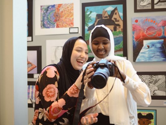 Two young people looking at elements of a camera with a long lens in a room with the wall behind them covered with artwork.