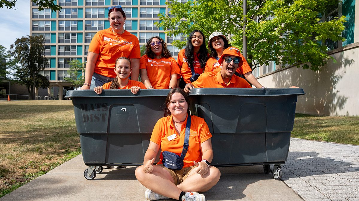 Seven people in matching orange shirts pose outdoors around a large black bin, with six people inside of it and one person sitting on the ground in front. A building, sidewalk and trees can be seen in the background.