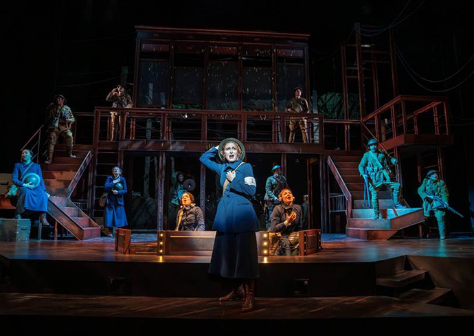 Stage performance featuring multiple actors in historical or military-themed period costumes, set on a two-level structure with stairs and various props