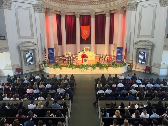 A large auditorium filled with seated attendees facing a stage where a speaker stands at a podium. Behind the speaker are several individuals, with red curtains and an orange banner featuring an emblem as the backdrop.