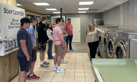 Group of people standing in a clean, well-lit laundromat with tiled flooring. One person is pointing at a washing machine, possibly explaining something to the group. A 'SOAP CENTER' sign on the wall reads 'SOAP BLEACH SOFTENER – $1.25 each,' and multiple washing machines are lined up along the wall.