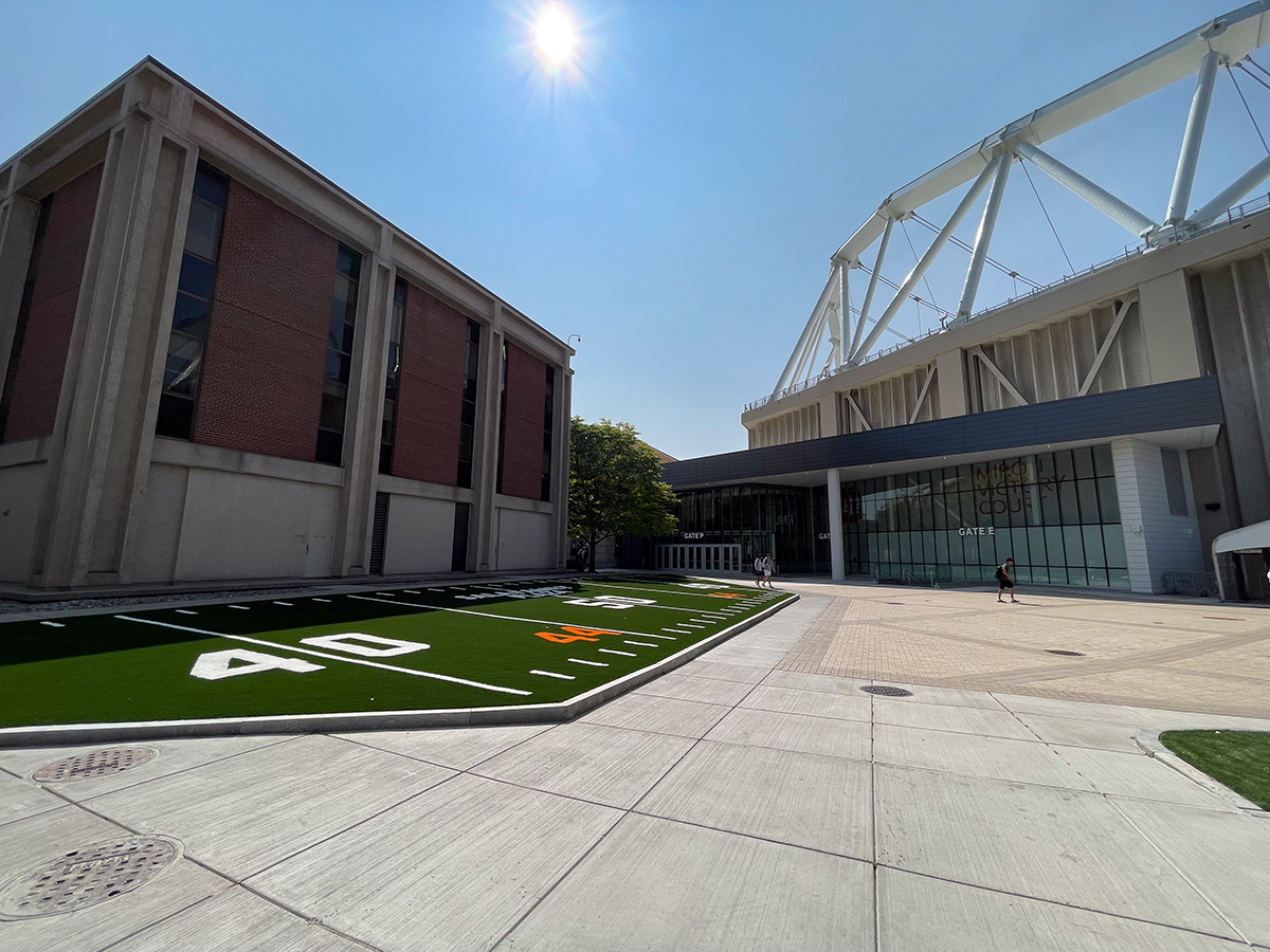 Outdoor scene featuring a small artificial turf area marked with '40', flanked by a brick building and a modern glass structure under a clear, sunny sky.