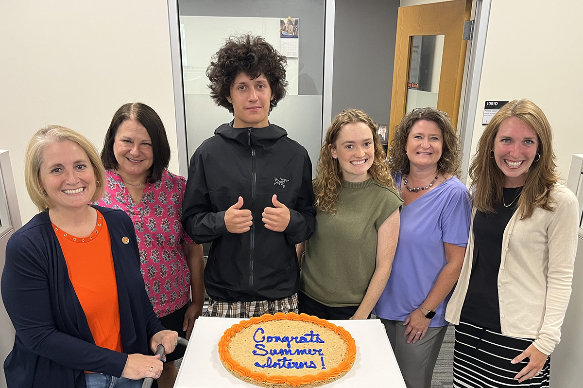 Six people standing behind a table with a cookie cake that reads 'Congrats Summer Interns!' in an office setting