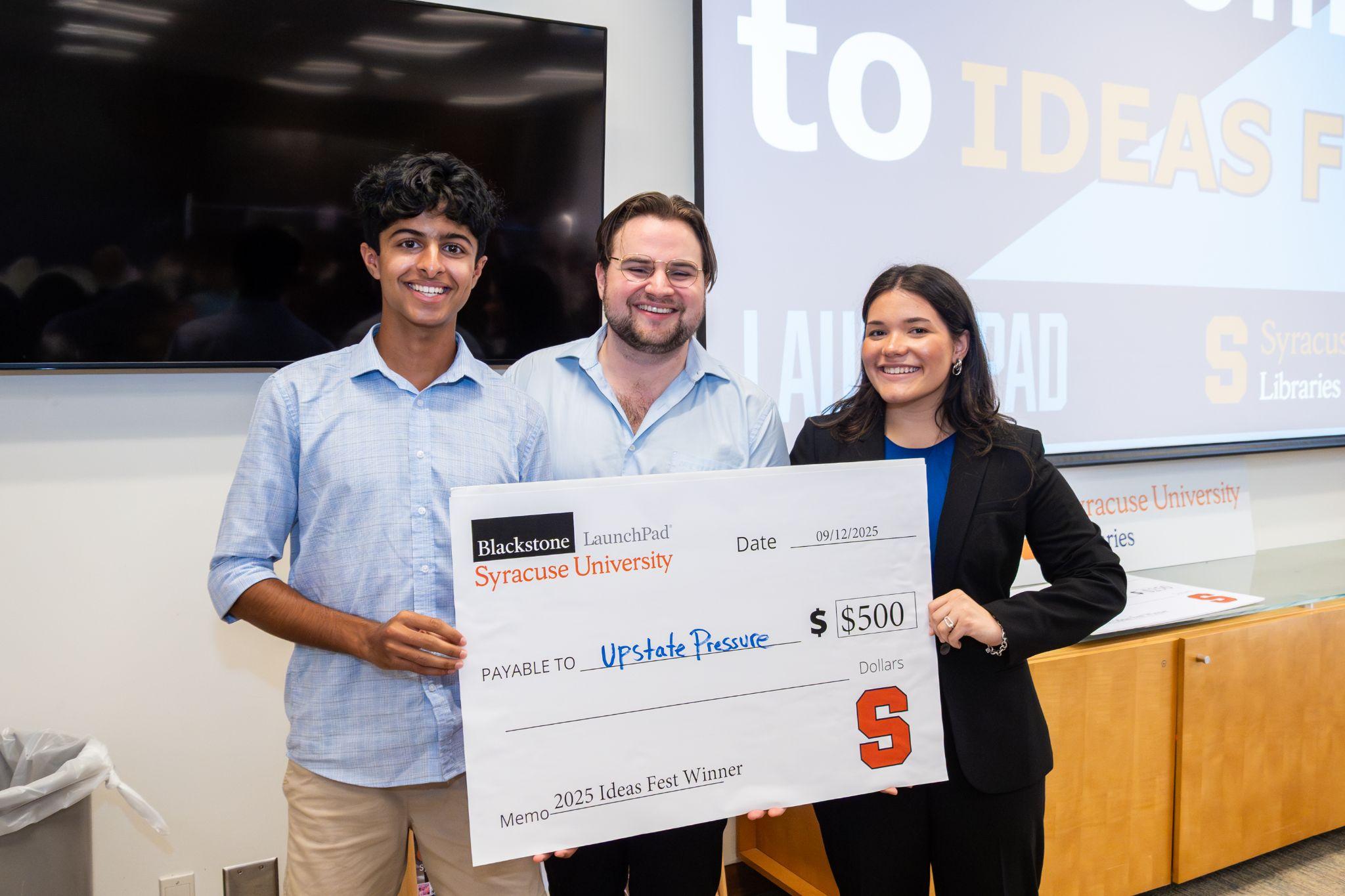Three people pose with a large $500 check from Blackstone LaunchPad at Syracuse University, awarded to "Upstate Rescue" as the 2025 Ideas Fest Winner, with a projection screen visible in the background.
