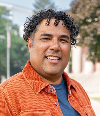 Person with curly hair in an orange corduroy jacket and blue shirt, standing outdoors with trees and a building in the background