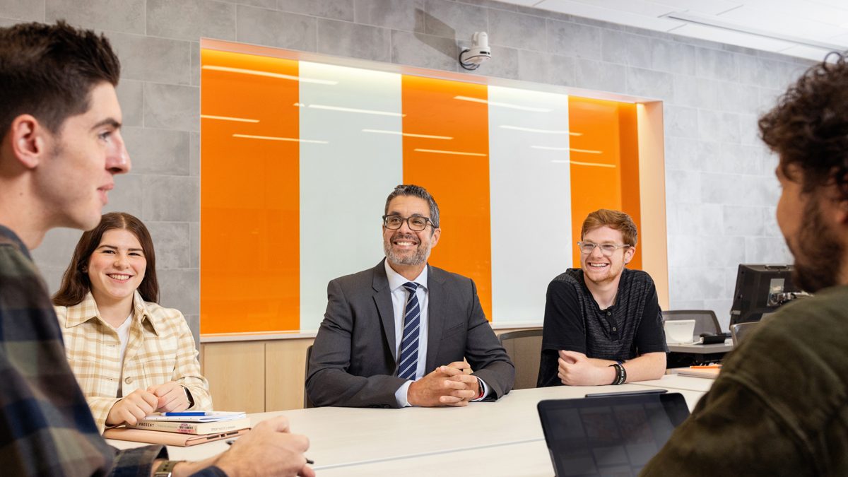 Five people, including a suited man and casually dressed individuals, sit around a table in a bright, modern room with orange and white wall panels, engaged in a collaborative discussion with notebooks and a tablet.