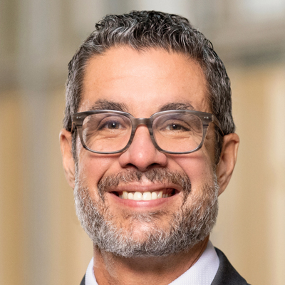 Close-up portrait of a smiling middle-aged man with short curly hair, salt-and-pepper beard, rectangular glasses, and a suit and tie, set against a softly blurred indoor background.