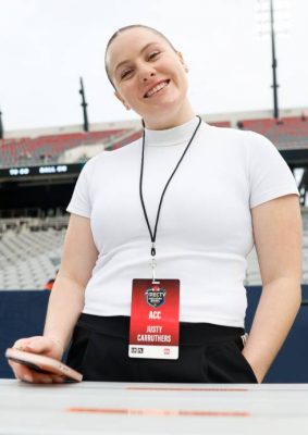 Person wearing a white turtleneck and black pants stands in a stadium with a "DIRECTV ACC" ID badge, holding a smartphone with empty seats and a field in the background