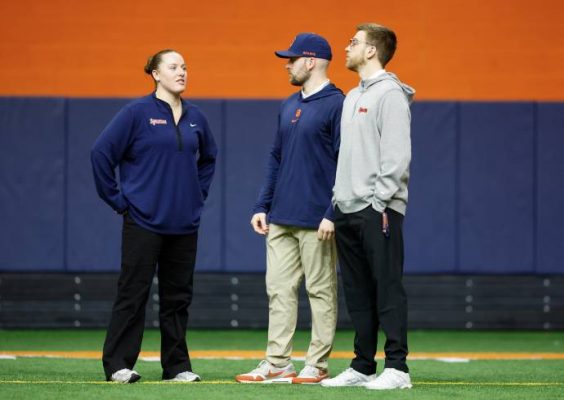 Three people standing on an indoor sports field with a blue and orange backdrop, dressed in casual athletic wear including jackets, hoodies and sneakers.