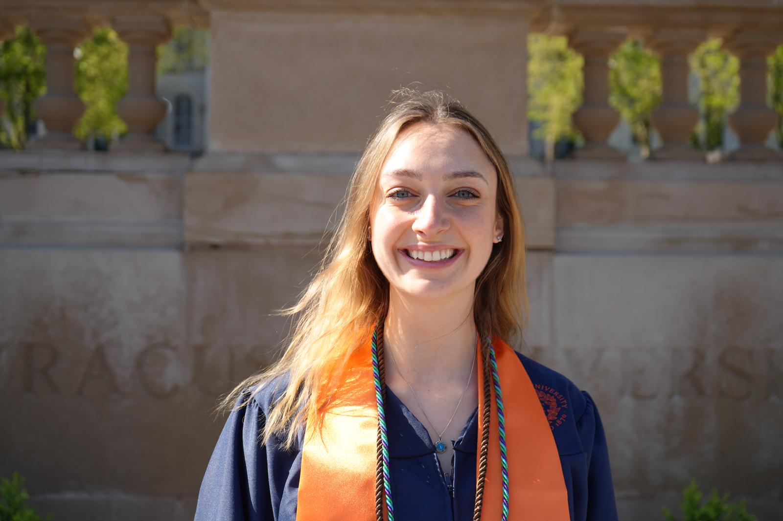 A smiling person with long blonde hair wearing a dark graduation gown with an orange and green honor cord, photographed outdoors with a building in the background.