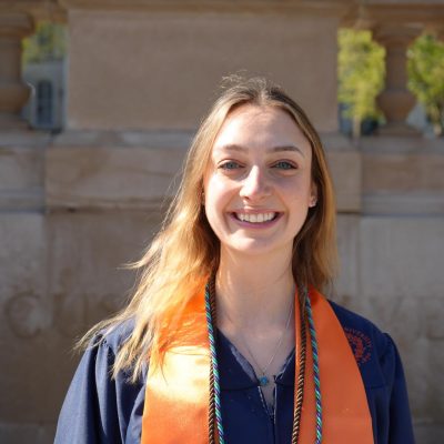 A smiling person with long hair wearing a dark graduation gown with an orange and green honor cord, photographed outdoors