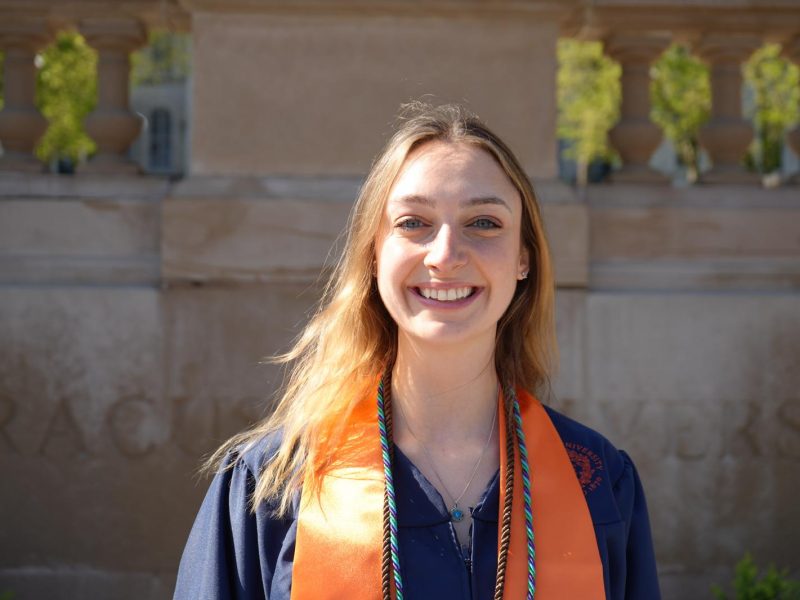 A smiling person with long hair wearing a dark graduation gown with an orange and green honor cord, photographed outdoors