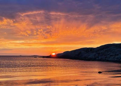 Golden sunset over calm water with dramatic orange and purple clouds reflected on the surface, silhouetted rocky coastline in background.