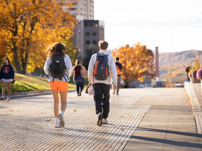 Students walking along a tree-lined campus pathway during fall, with vibrant orange and yellow foliage and scenic hills in the background