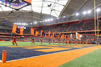 Cheerleaders running across a football field in a domed stadium, each holding an orange flag with a blue letter that collectively spells 'SYRACUSE'