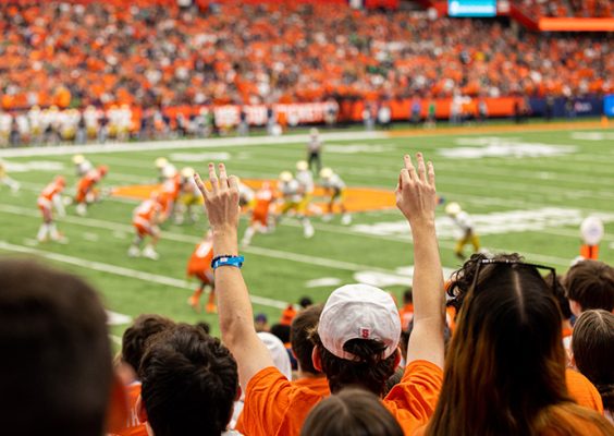 Fans in orange cheer from stadium stands during a football game, with one fan in the foreground raising both arms, three fingers extended on each hand.