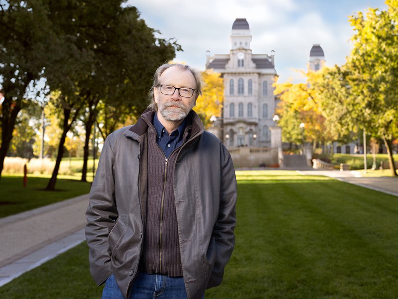 a man in a grey jacket stands on campus with Hall of Languages and fall foliage in the background