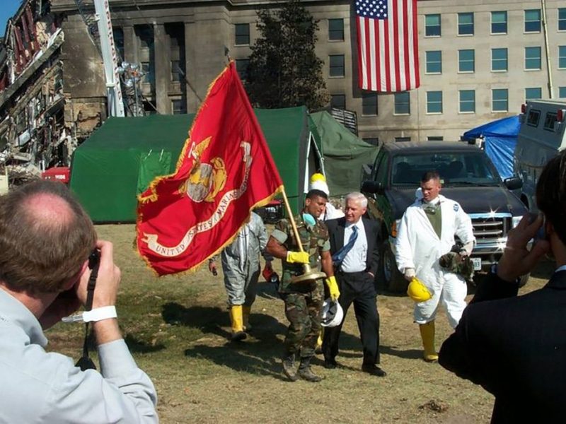 Military personnel and hazmat-suited individuals gather outside a damaged building, with a U.S. Marine Corps flag prominently displayed and an American flag hanging in the background; emergency vehicles and tents are present