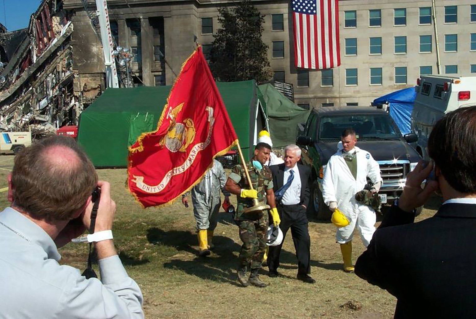 Military personnel and hazmat-suited individuals gather outside a damaged building, with a U.S. Marine Corps flag prominently displayed and an American flag hanging in the background; emergency vehicles and tents are present.