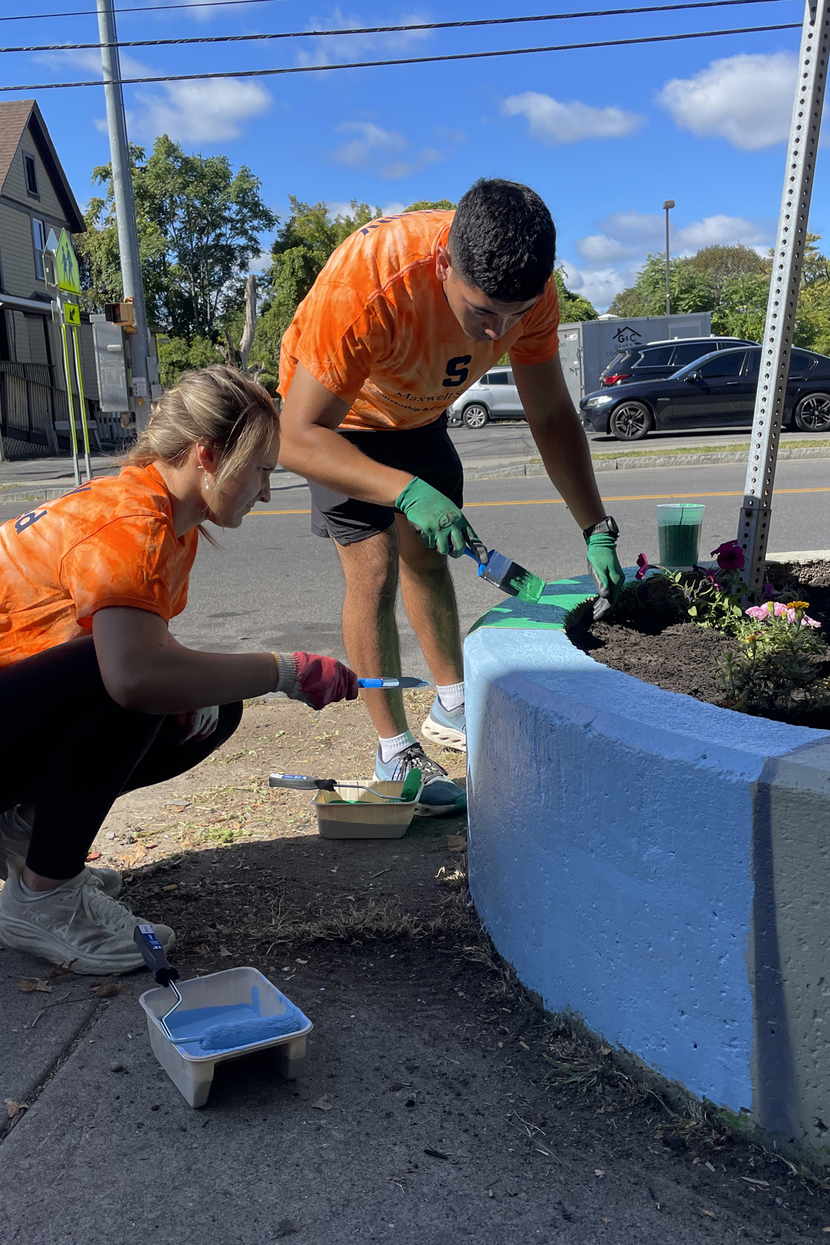Two volunteers in orange shirts painting a blue concrete planter and planting flowers on a city street, with paint supplies nearby and residential buildings in the background.