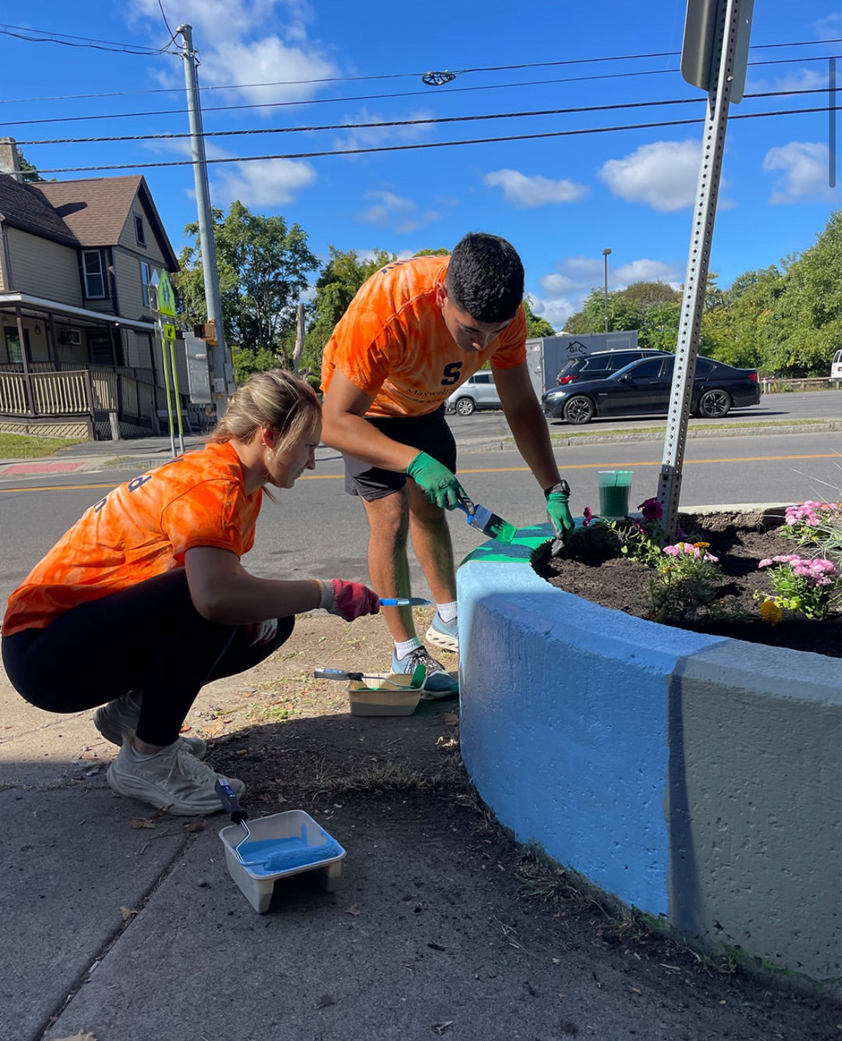 Two volunteers in orange shirts painting a blue concrete planter and planting flowers on a city street, with paint supplies nearby and residential buildings in the background.