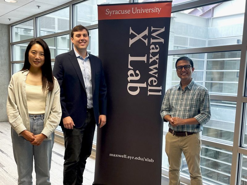 Three people stand beside a “Maxwell X Lab” banner in a sunlit hallway