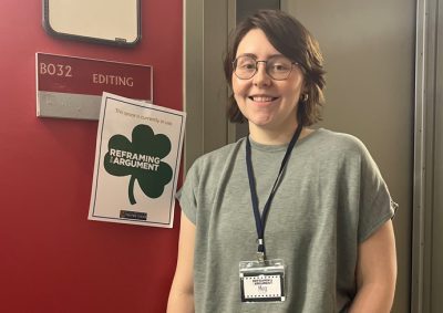 A person with short brown hair and glasses stands smiling in front of a red door marked "B032 EDITING." They're wearing a gray t-shirt and jeans with a name tag lanyard around their neck.