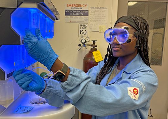 A woman wearing safety goggles, blue gloves, and a light blue lab coat works with a glowing blue laboratory instrument in a research lab.