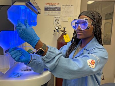 A woman wearing safety goggles, blue gloves, and a light blue lab coat works with a glowing blue laboratory instrument in a research lab