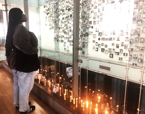 Person standing before a glass wall filled with small black-and-white photographs, with lit candles along the bottom edge, suggesting a memorial