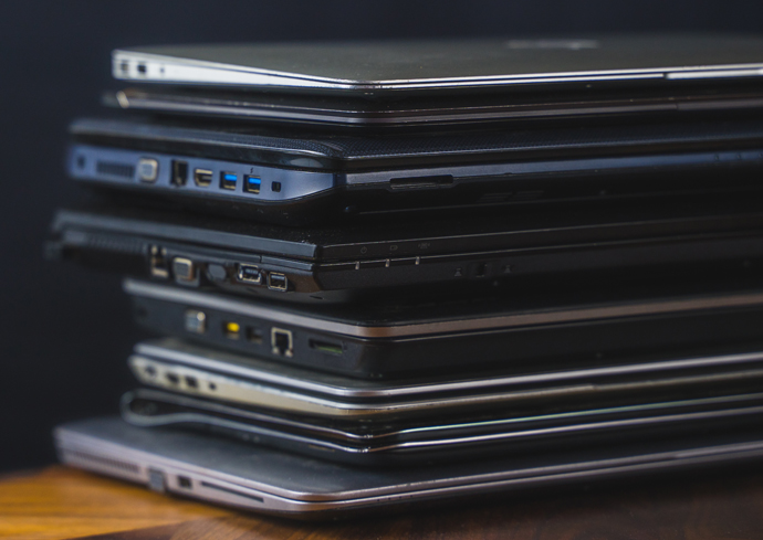 A stack of several closed laptops piled on top of each other, showing their side profiles with various ports like USB, HDMI, and charging ports visible along the edges.