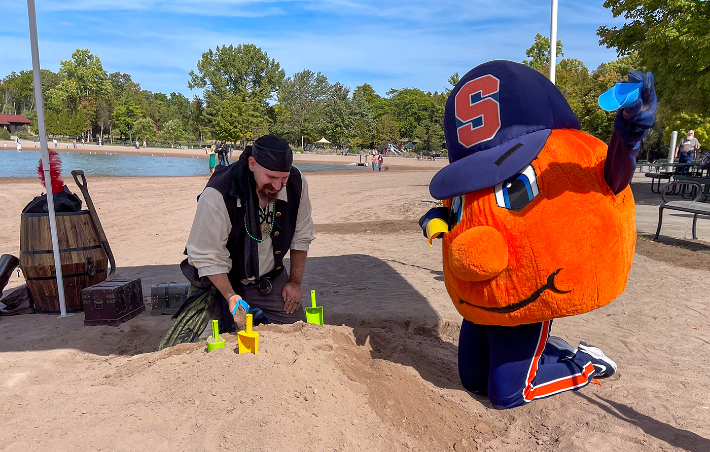 Person in pirate costume and orange mascot with 'S' cap build sandcastles on a beach using colorful molds and a blue shovel, with trees and water in the background.
