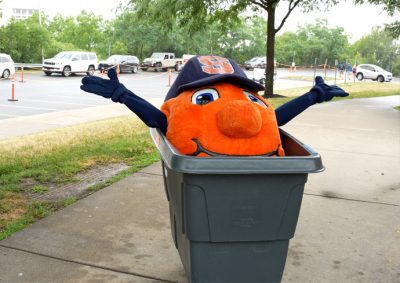 Otto the Orange, smiling and waving from a trash bin, outdoors near a parking lot.