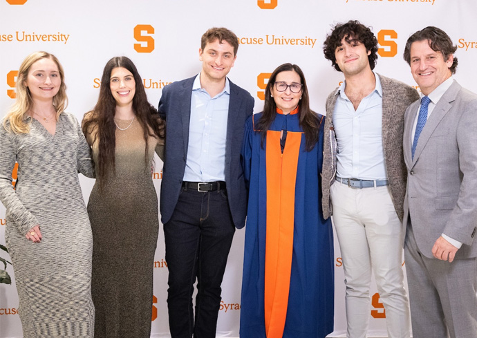 Trustee Jeannine Lostritto, in Commencement robe, with her husband and children standing in front of a Syracuse University step and repeat