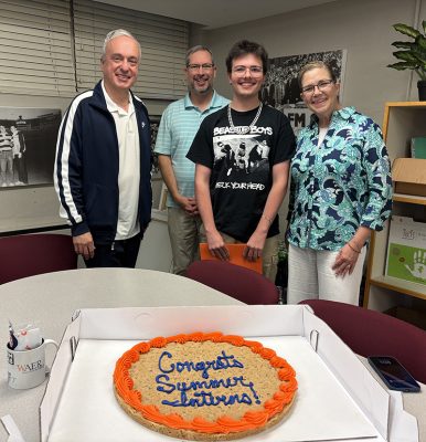 Four people behind a table with a cookie cake that reads 'Congrats Summer Interns!' alongside a WAER mug and smartphone, in an office setting