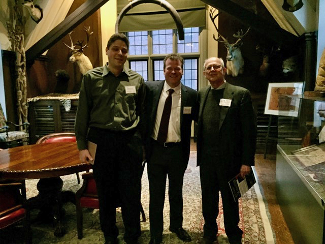 Three men posing together in an ornate indoor setting with mounted deer heads on the walls, decorative wooden furniture, and large windows. The men are wearing business casual attire with name tags, suggesting this is a professional or conference event.
