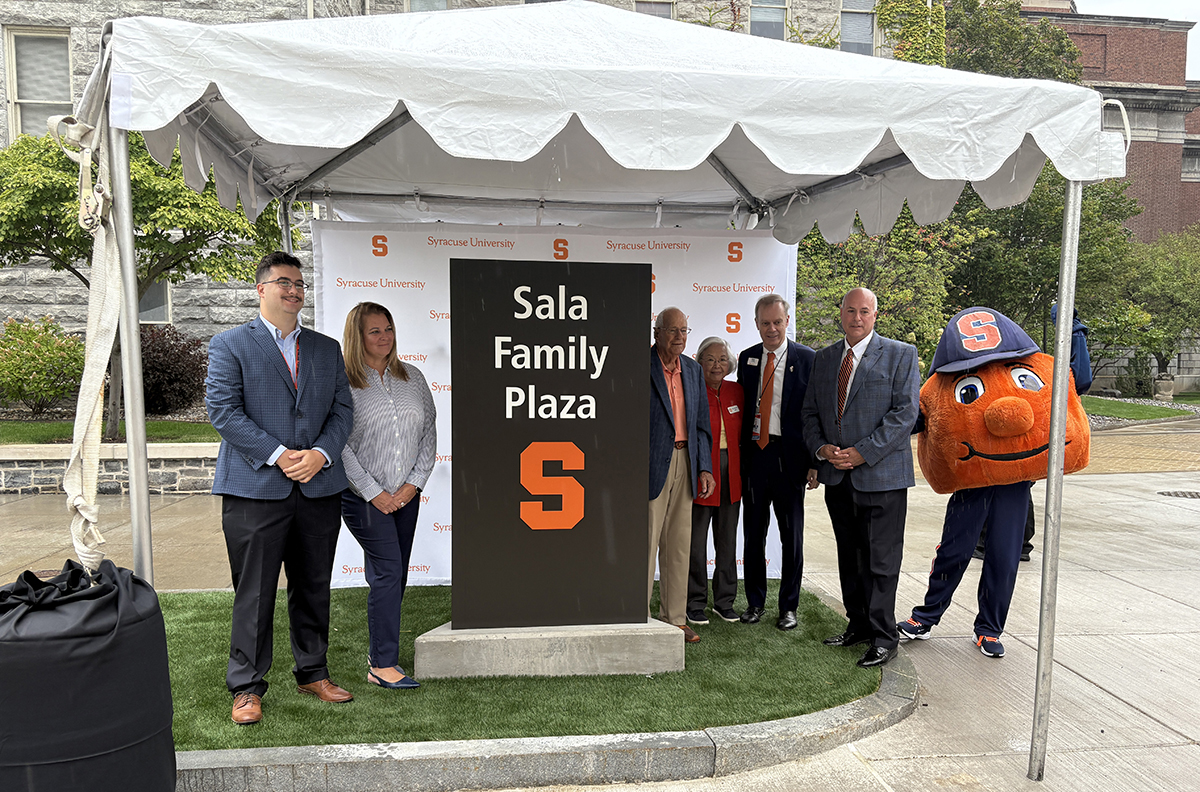 Group of six people and the Syracuse University mascot Otto the Orange pose beside a monument sign reading "Sala Family Plaza" with Syracuse University's orange "S" logo at a dedication ceremony under a white tent.