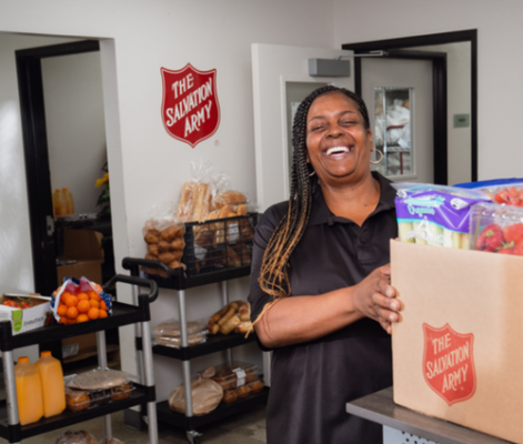 A smiling person stands in a Salvation Army food pantry, holding a paper bag with the organization's red shield logo. Behind her are shelves stocked with bread, fresh produce including oranges, and other food items in an organized distribution area.