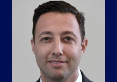 A professional headshot of a man with dark hair wearing a dark business suit, white dress shirt, and gray plaid tie, smiling slightly at the camera against a neutral gray background.